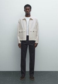 Beige jacket with two front pockets and buttons, over a white shirt; paired with dark denim pants and brown shoes, set against a light background.