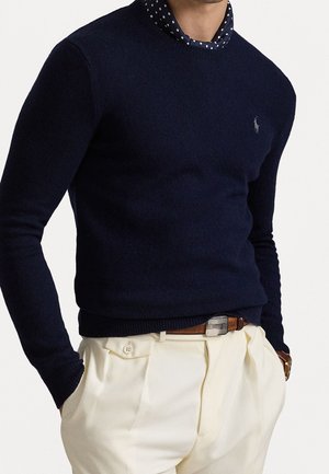 Mand iført navyblå crewneck-trøje over navy skjorte med hvide prikker, stukket ned i højtaljede cremefarvede bukser med brun bælte.