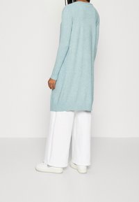 Jasnoniebieski sweter z długim rękawem o luźnym kroju, z prążkowanymi mankietami i bocznymi rozporkami, zestawiony z szerokimi białymi spodniami i białymi sneakersami.