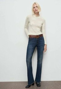 Suéter blanco de punto con canalé y mangas largas; jeans de campana azul oscuro con dobladillos deshilachados; cinturón marrón con hebilla metálica redonda; botas negras hasta el tobillo.