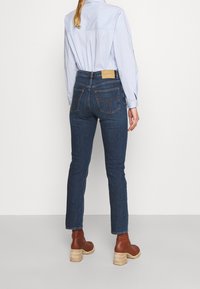 Mujer vestida con jeans ajustados de color azul oscuro, camiseta de manga larga de color azul claro y botas de tacón marrones, de pie frente a un fondo blanco liso.