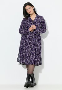 Paarse shirtjurk met een bloemenpatroon in roze en wit. Voorzien van lange mouwen en een lengte tot halverwege de kuiten, gecombineerd met zwarte veterschoenen.