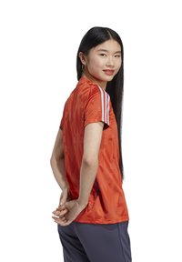 T-shirt de sport orange avec un design texturé, bandes d'accent bleu sur les manches, et manches courtes. Porté avec un pantalon de sport gris foncé.