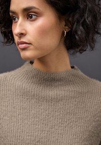 Beige ribbed knit sweater dengan leher tinggi. Model menunjukkan tekstur kain dan rambut ikal alami. Aksen anting bulat emas sederhana.