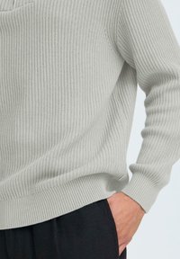 Hellgrauer Strickpullover mit gerippter Textur, runder Ausschnitt und langen Ärmeln, kombiniert mit schwarzen Hosen.