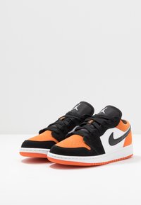 Sneaker neri, bianchi e arancioni realizzati in materiali scamosciati e in rete. Presentano punta perforata, lacci neri e suole curve per una migliore trazione.