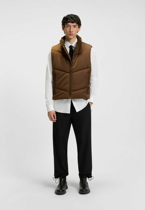 Bruine gewatteerde vest met hoge kraag, voorrits en zijzakken, gedragen over een witte shirt en zwarte broek. Glanzende textuur.