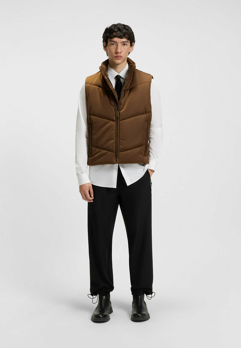 Gilet imbottito marrone con colletto alto, cerniera anteriore e tasche laterali, indossato su una camicia bianca e pantaloni neri. Texture lucida.