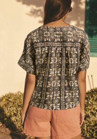 Blouse à manches courtes présentant un fond sombre avec un motif floral et géométrique crème. Associée à un short rose clair.