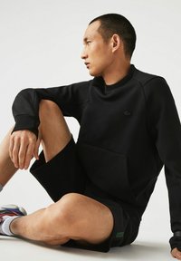 Sudadera negra con cuello alto, bolsillo frontal y puños acanalados. Combinada con pantalones cortos negros. Textura suave con un diseño minimalista.