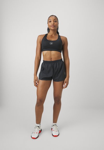 Sujetador deportivo negro con logo, combinado con pantalones cortos negros con cinturilla elástica. El modelo lleva zapatillas deportivas blancas y rojas.