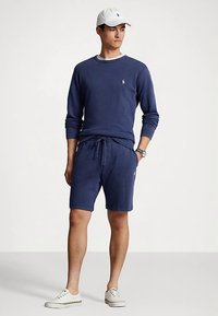 Φούτερ σε ναυτικό μπλε με στρογγυλή λαιμόκοψη και λευκό περιθώριο, συνδυασμένο με ναυτικά σορτς με ελαστικό κορδόνι. Λευκό καπέλο και παπούτσια.