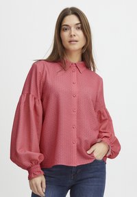 Rote Bluse mit Muster, Knopfleiste, großen Ballonärmeln und elastischen Bündchen. Über dunklen Jeans getragen, zeigt sie eine lässige Passform.