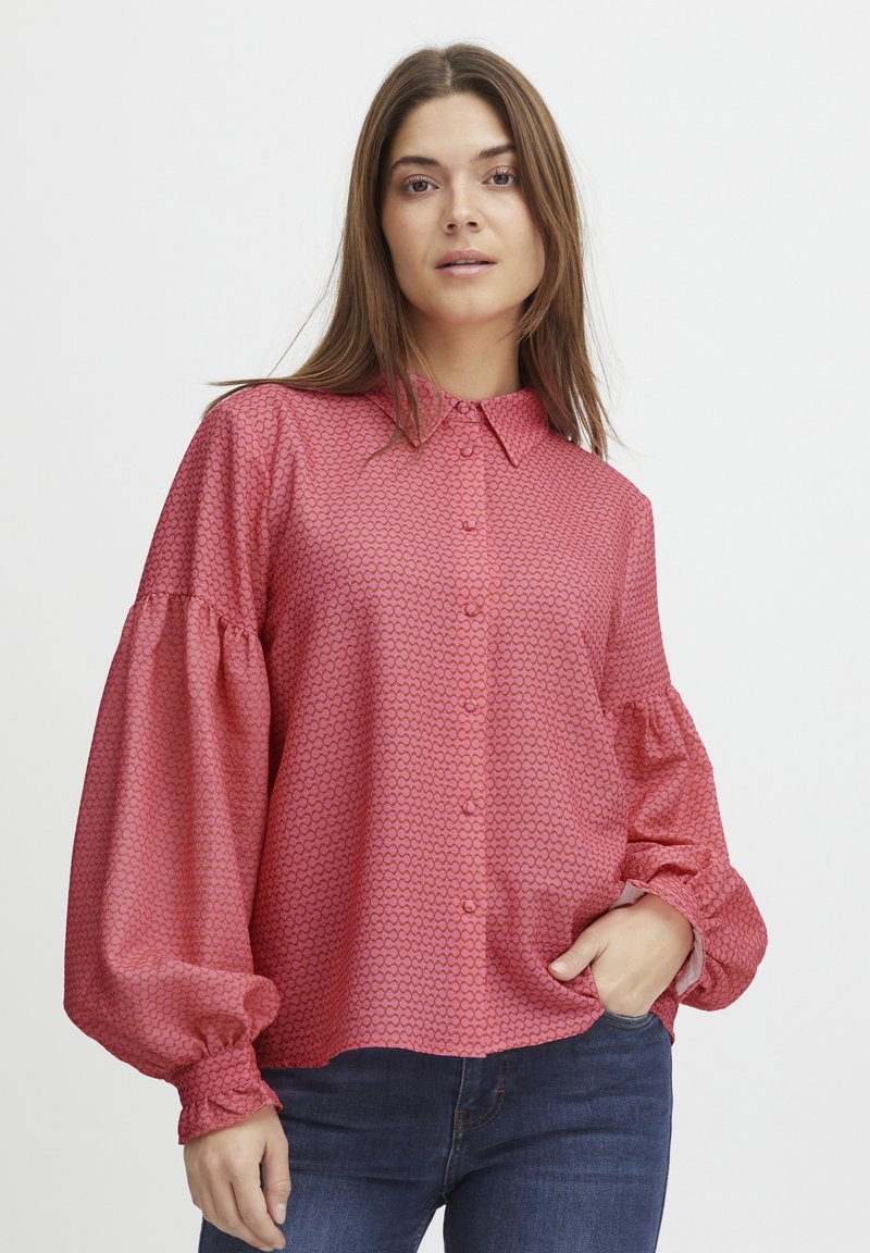 Blusa rossa a fantasia con bottone frontale, ampie maniche a palloncino e polsini elasticizzati. Indossata sopra jeans scuri, mostra un fit rilassato.