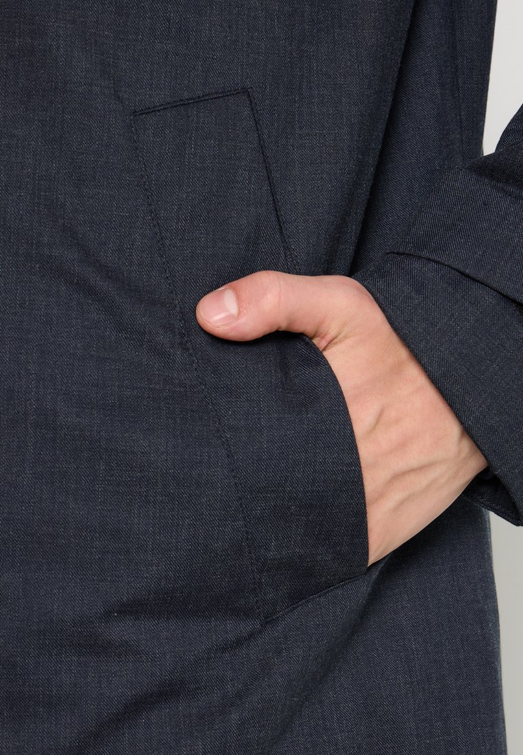 Veste bleu marine en tissu texturé, dotée d'une poche latérale. Main insérée dans la poche, mettant en avant un design minimaliste.