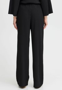 Pantaloni neri a gamba larga con texture a costine verticali, dotati di una vita elastica e una finitura in tessuto liscia. Le gambe scendono dritte fino al pavimento.