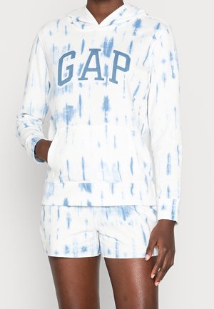 Osoba nosząca biało-niebieską bluzę z kapturem tie-dye z napisem „GAP” i pasujące szorty, ręka w kieszeni bluzy, stojąca na jednolitym tle.