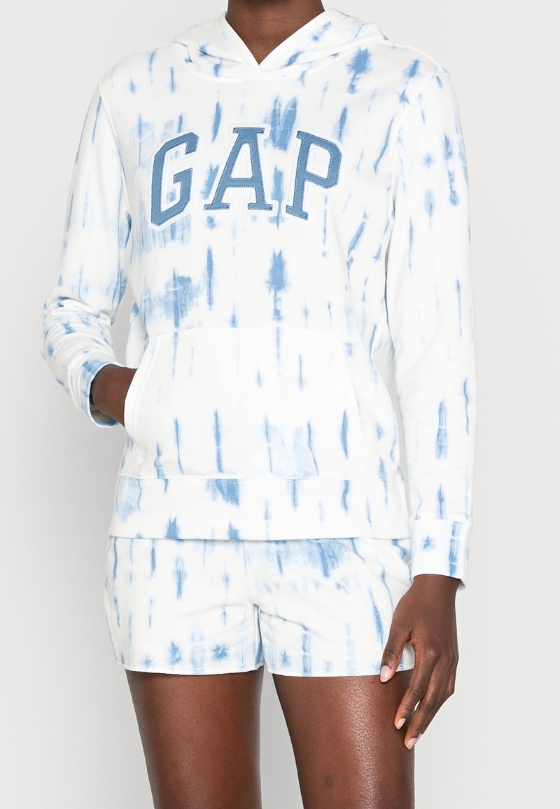 Personne portant un sweat à capuche tie-dye blanc et bleu avec l’inscription « GAP » et un short assorti, main dans la poche du sweat, debout devant un fond uni.