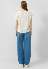 Mujer de pie de espaldas, vestida con una blusa blanca de manga corta con ojales, pantalones vaqueros azules de pierna ancha y zapatos blancos de punta cuadrada sobre un fondo liso.