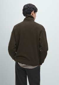 Maglione dolcevita marrone scuro in tessuto a maglia strutturato, con vestibilità comoda e polsini e orlo a coste. Indossato sopra una camicia grigio chiaro.