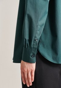 Mörkgrön skjorta med knappar framtill och en åtsittande manschett, med en subtil struktur och elegant design. Inkluderar mörka knappar vid manschetten.