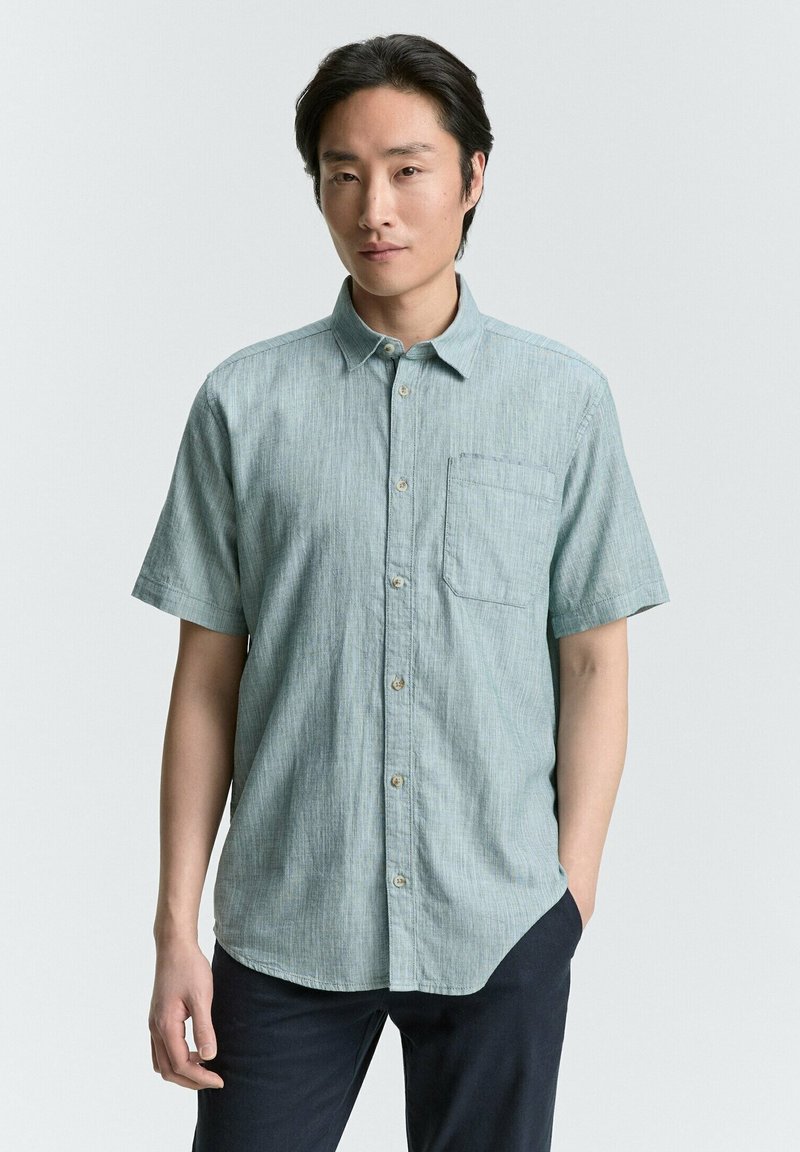 Chemise à manches courtes bleu clair avec un col boutonné, une poche avant et une finition en tissu texturé. Présente un ourlet droit et des fermetures à boutons.