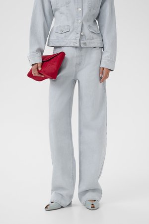 Kobieta w jasnoniebieskiej kurtce dżinsowej i szerokich dżinsach, trzymająca czerwoną kopertówkę, w jasnoniebieskich sandałach na obcasie z odkrytymi palcami.