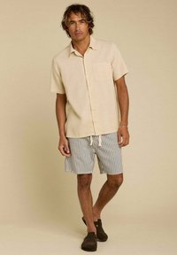 Chemise beige à manches courtes avec un bouton et une poche, accompagnée de shorts rayés aux couleurs claires et foncées. Chaussures brunes à enfiler.
