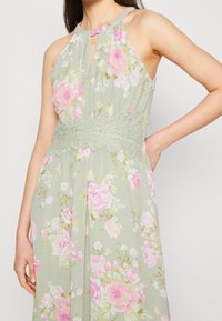 Kukkakuvioinen vaaleanvihreä mekko, jossa on vaaleanpunaisia ruusuja, pleatattu vartalo, avainaukko-yleinen yksityiskohta ja monimutkainen pitsivyötärö.