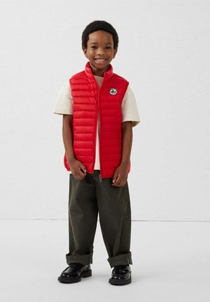 Rode gewatteerde bodywarmer met een hoge kraag en logopatch, gedragen over een beige t-shirt. Gecombineerd met olijfgroene broek en zwarte schoenen.