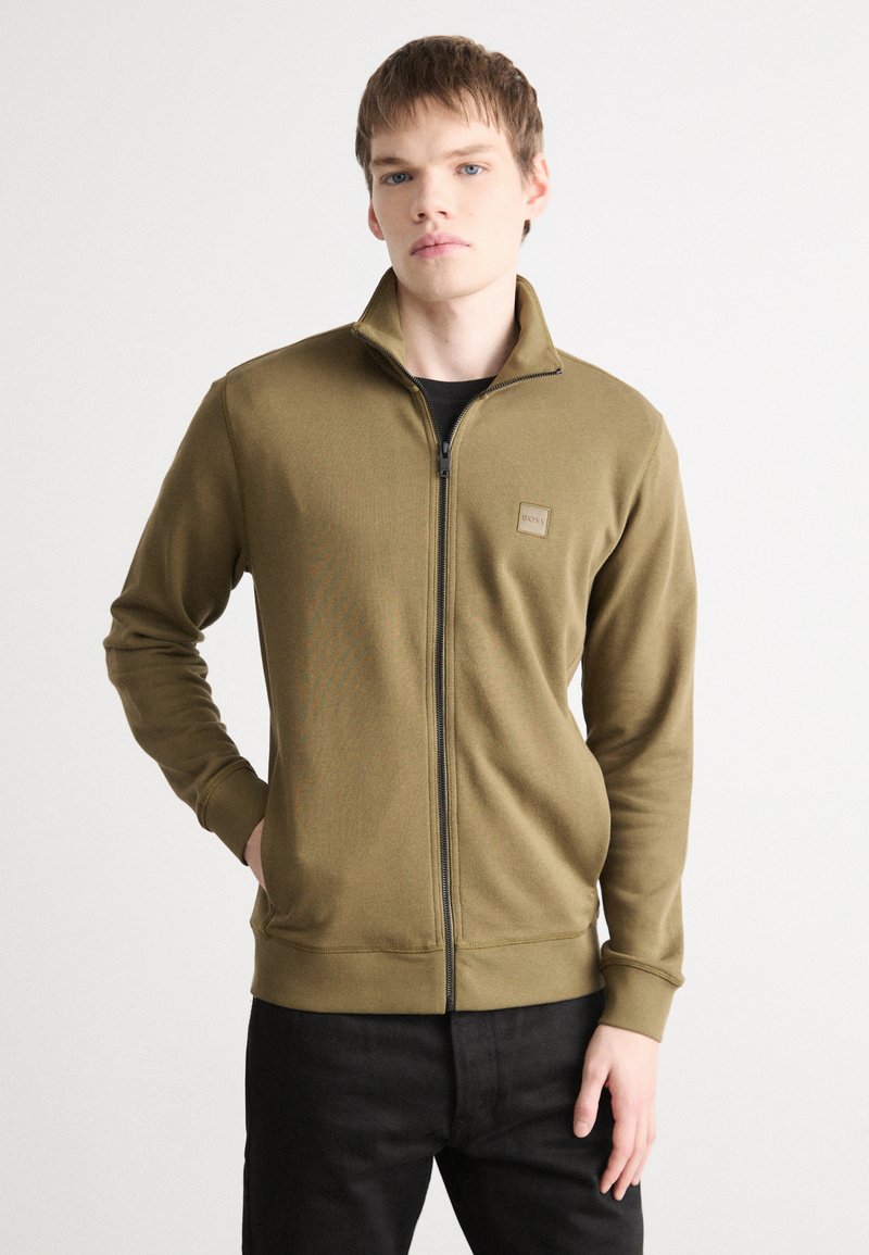 Oliven grønn zip-up jakke med krage, med teksturert stoff, ribbestrikkede mansjetter og et lite logo-patch på brystet.
