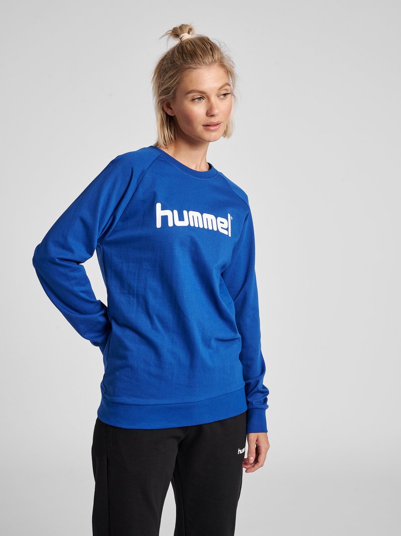 Kvinde med blondt hår iført en blå "hummel" sweatshirt og sorte bukser, stående med den ene hånd på hoften og kigger let til siden.