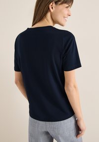 Femme souriante, portant un t-shirt bleu marine uni à manches courtes et un pantalon gris clair, debout, de dos en grande partie tournée vers la caméra.