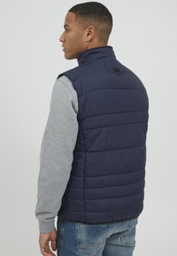 Námornícky modrá prešívaná vesta s vysokým golierom a sivým dlhým rukávom pod ňou. Na zadnej strane obsahuje značkovú nálepku.