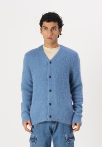 Μπλε, χνουδωτό καζάκι από μαλακό υλικό με V ντεκολτέ, μακριά μανίκια και έξι μαύρα κουμπιά μπροστά. Οι άκρες είναι ελαφρώς φθαρμένες.