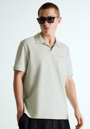 Junger Mann trägt schwarze Sonnenbrille, ein hellbeiges kurzärmeliges Poloshirt mit kleinem Logo und schwarze Hose, steht vor einem schlichten Hintergrund.