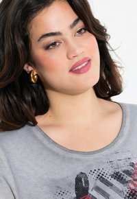 T-shirt gris avec un col rond et un design graphique texturé en rouge et noir. Le modèle porte des boucles d'oreilles en or. Finition en tissu lisse.