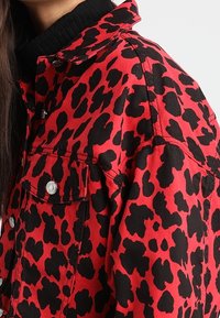 Röd denimjacka med ett svart leopardmönster, med krage, tryckknappar och en mjuk textur.