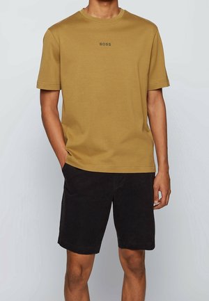 Mężczyzna w żółtej koszulce BOSS z okrągłym dekoltem i czarnych krótkich spodenkach do kolan, stojący z jedną ręką w kieszeni na tle jednolitego tła.