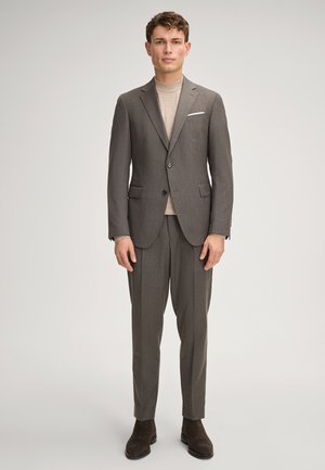 Mężczyzna ubrany w skrojony na miarę brązowy garnitur z dwuguzikową marynarką, beżowym golfem, brązowymi spodniami i ciemnobrązowymi botkami, stojący na neutralnym tle.