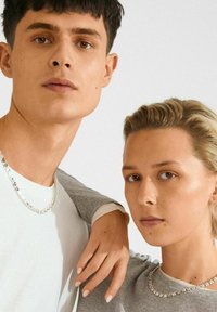 Joven hombre y mujer con collares de cadena plateados, vestimenta neutral y expresiones serias, frente a un fondo liso.