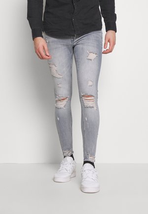 Homme portant un jean skinny déchiré gris clair, des baskets blanches, des chaussettes noires et une chemise noire à manches longues boutonnée, debout sur un fond blanc.