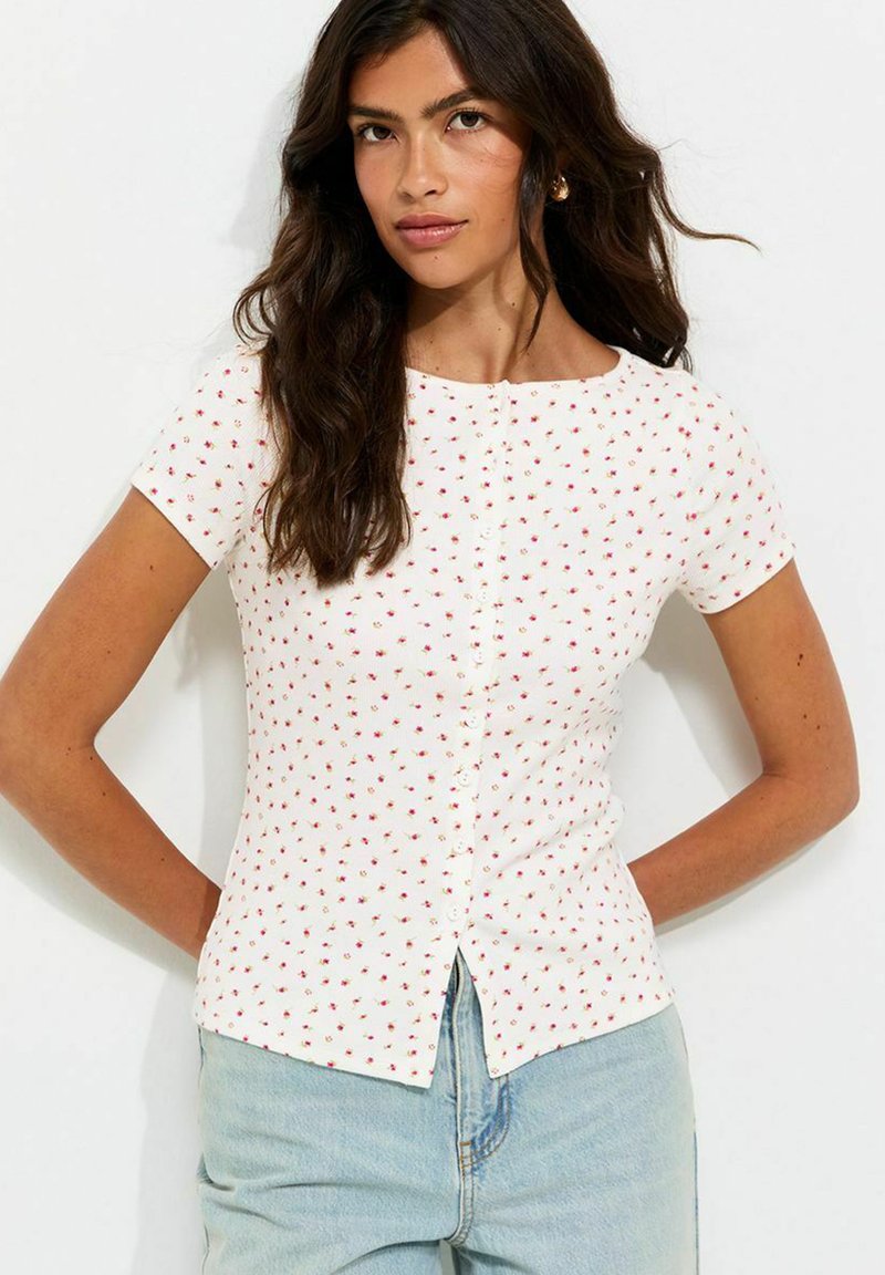 Mujer con cabello largo y oscuro que lleva una camiseta blanca de manga corta con pequeño estampado floral rojo y vaqueros azul claro, de pie contra un fondo blanco.