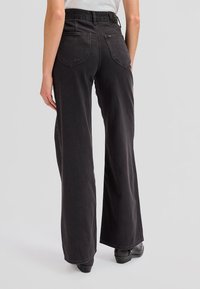 Woman wearing high-waisted, wide-leg black jeans with back pockets, paired with black ankle boots against a plain background.