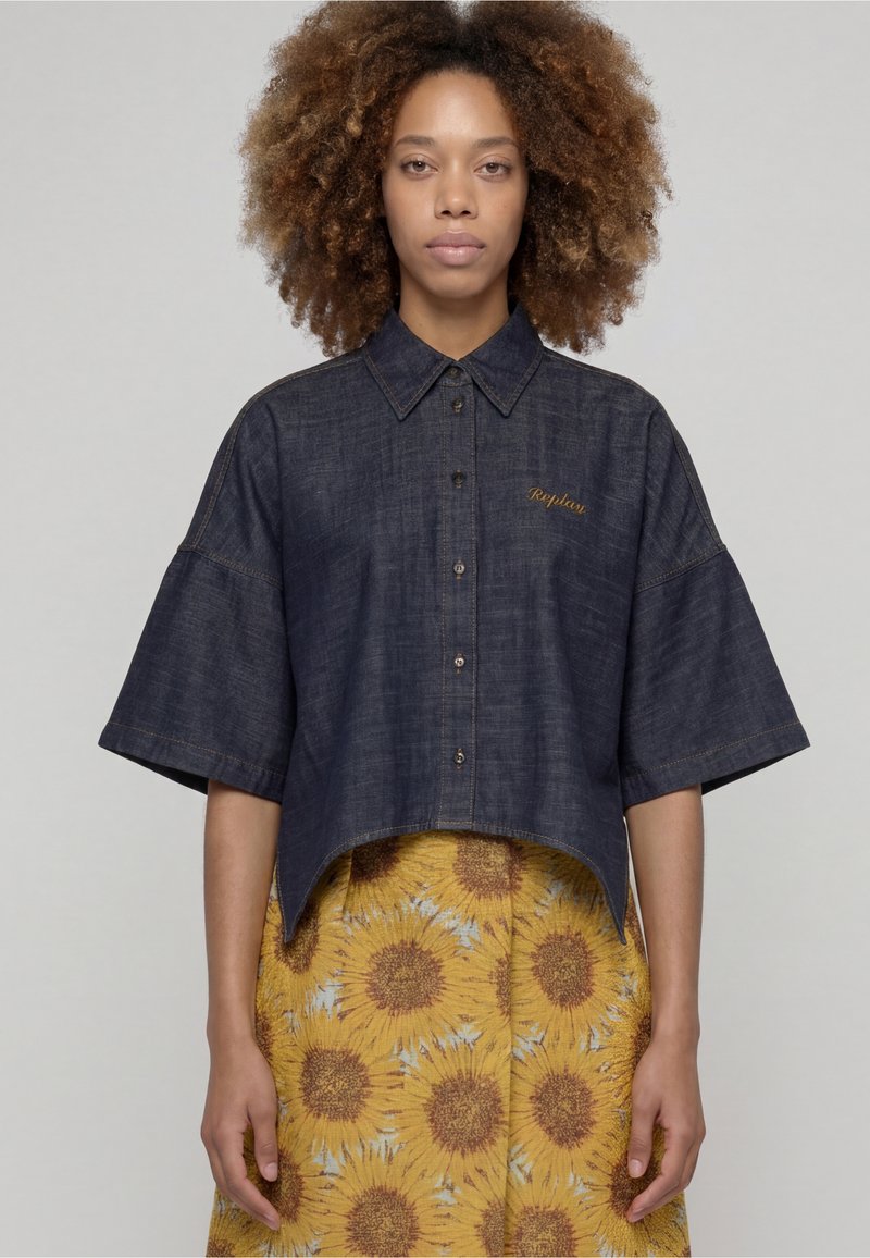 Personne aux cheveux bouclés porte une chemise en jean sombre ample à manches larges et une jupe jaune ornée d’un grand motif de tournesol, immobile.
