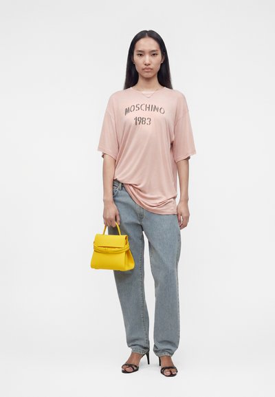 Femme portant un t-shirt rose ample Moschino 1983, jean bleu clair, talons noirs, tenant un petit sac à main jaune vif à ses côtés.