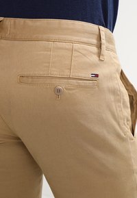 Kaki kalhoty z bavlny, s knoflíkovou zadní kapsou s kontrastním šitím a malou cedulkou s logem v červené, bílé a modré barvě.