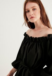 Svart off-shoulder klänning med volangkrage, 3/4 ärmar, elastisk midja och ett tygbälte. Slät textur. Enkel design.
