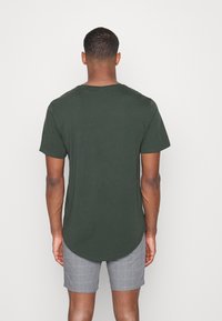 Donkergroen t-shirt met korte mouwen en een ronde hals, voorzien van een gebogen zoom en een zachte textuur. Gecombineerd met grijze geblokte shorts.
