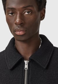 Zwarte zip-up jas van een getextureerd, fleece-achtig materiaal, met een kraag en zilveren rits hardware. Minimalistisch ontwerp, geen patronen.